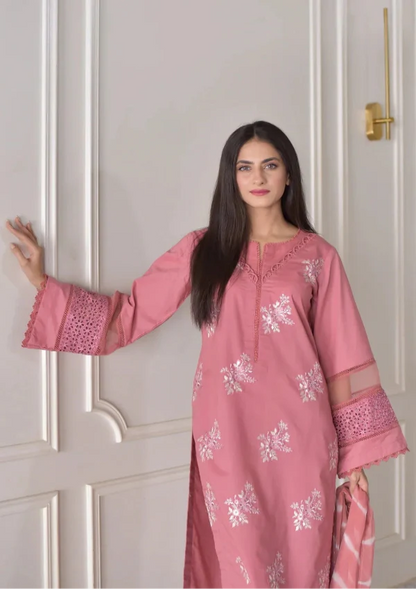 Woman wearing a pink traditional outfit with white patterns against a white paneled wall.